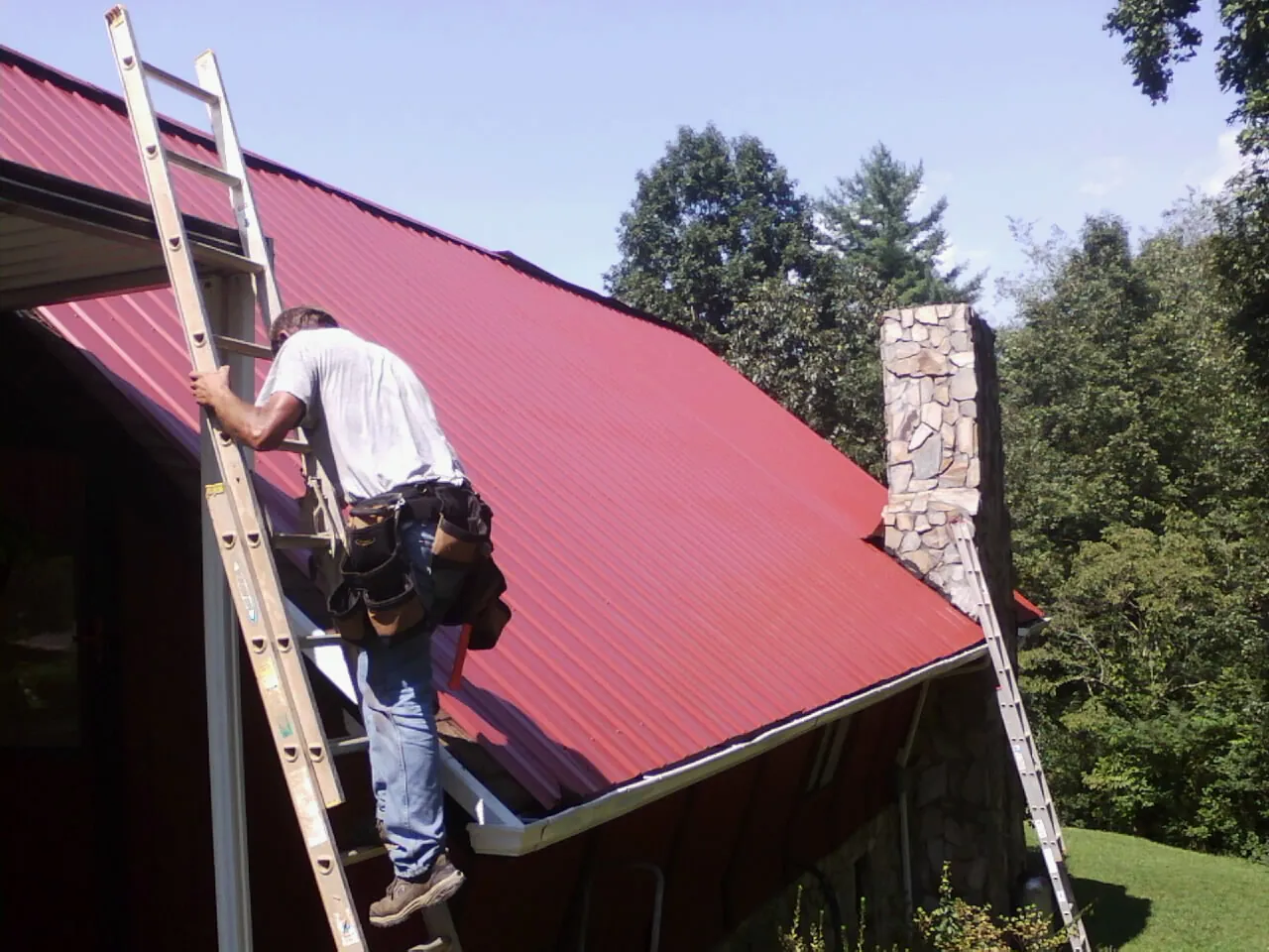 Roofer on a ladder beside a red metal roof and stone chimney — Soffit & Fascia Repair by Roofing Brothers in Belle Rose
